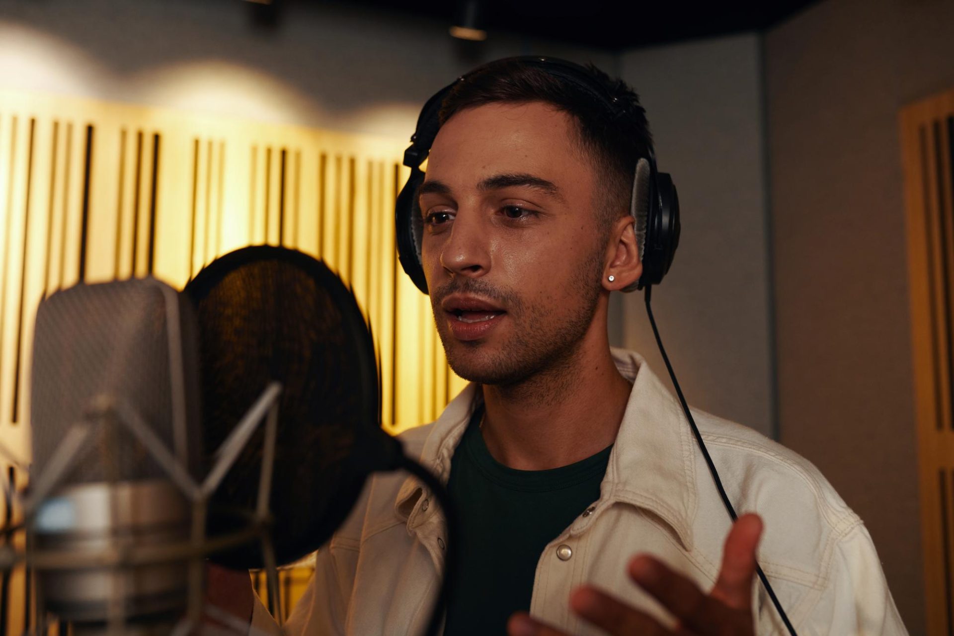 A young man wearing headphones stands in front of a microphone in a recording studio, speaking or singing with one hand raised, surrounded by acoustic panels.