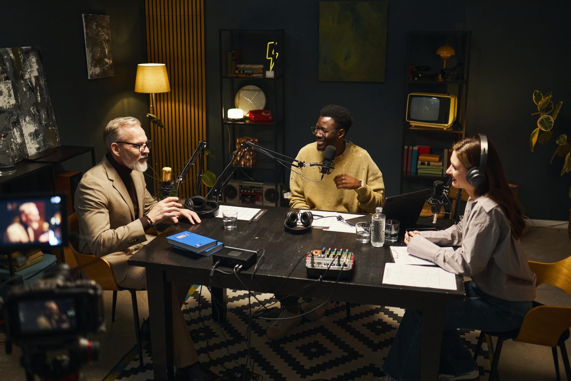Three people sit around a table with microphones and headphones, recording a podcast in a cozy, dimly lit studio with books, decorations, and recording equipment visible.