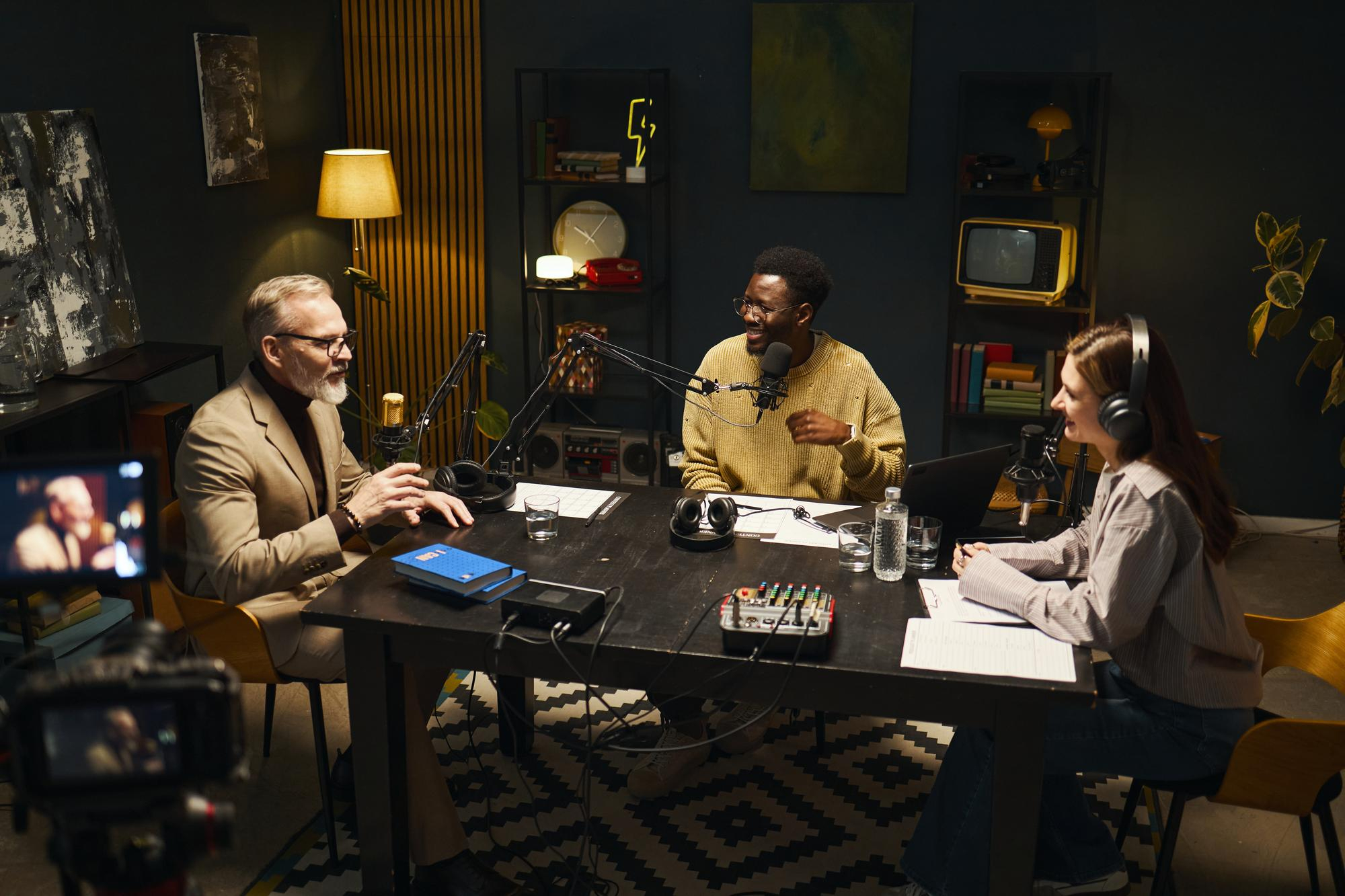 Three people sit around a table with microphones and headphones, recording a podcast in a cozy, dimly lit studio with books, decorations, and recording equipment visible.