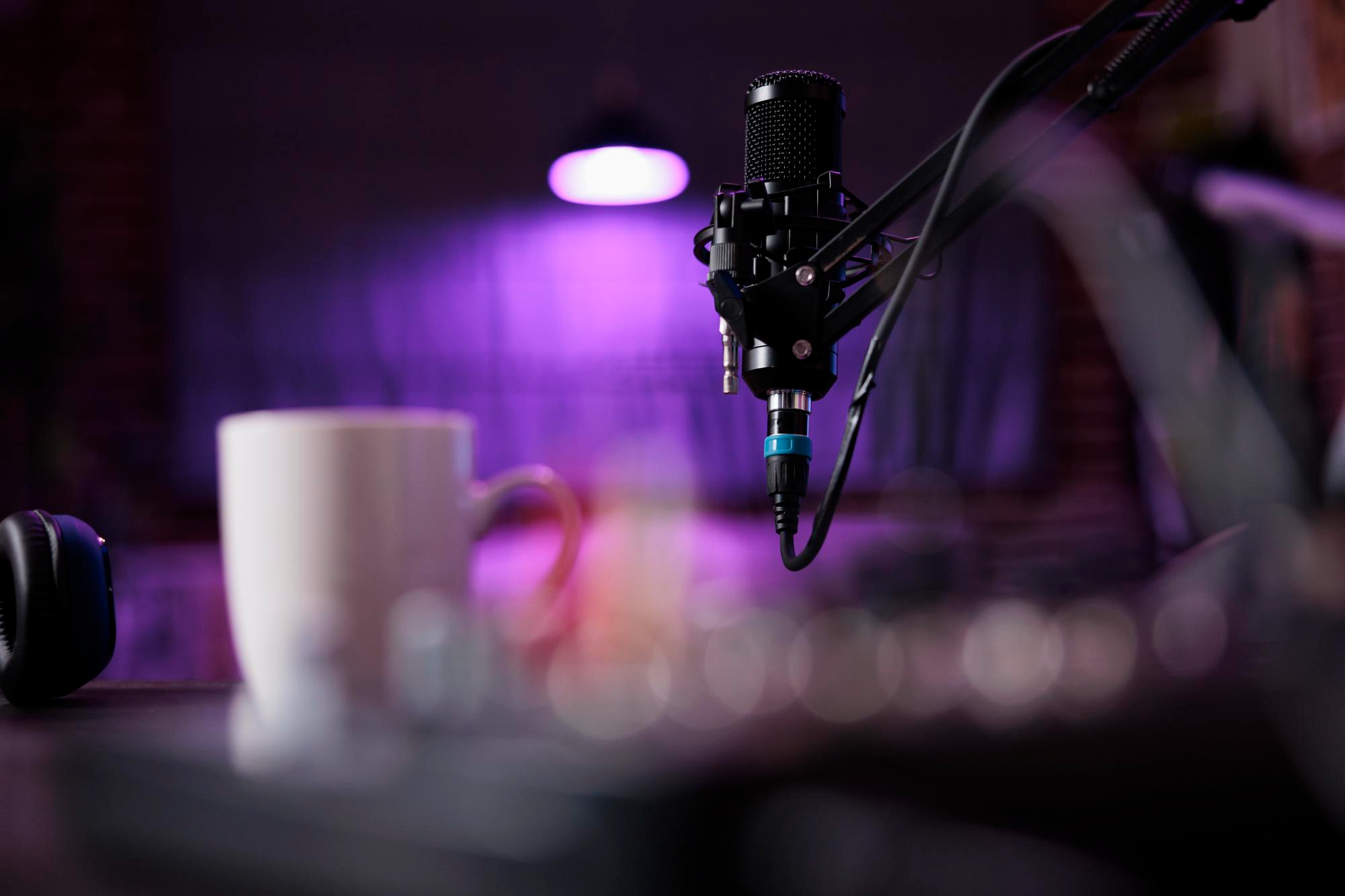 A close-up of a microphone on a stand, with a blurred white coffee mug and audio equipment in the foreground. The background is dimly lit with a purple light. podcast studio in Atlanta