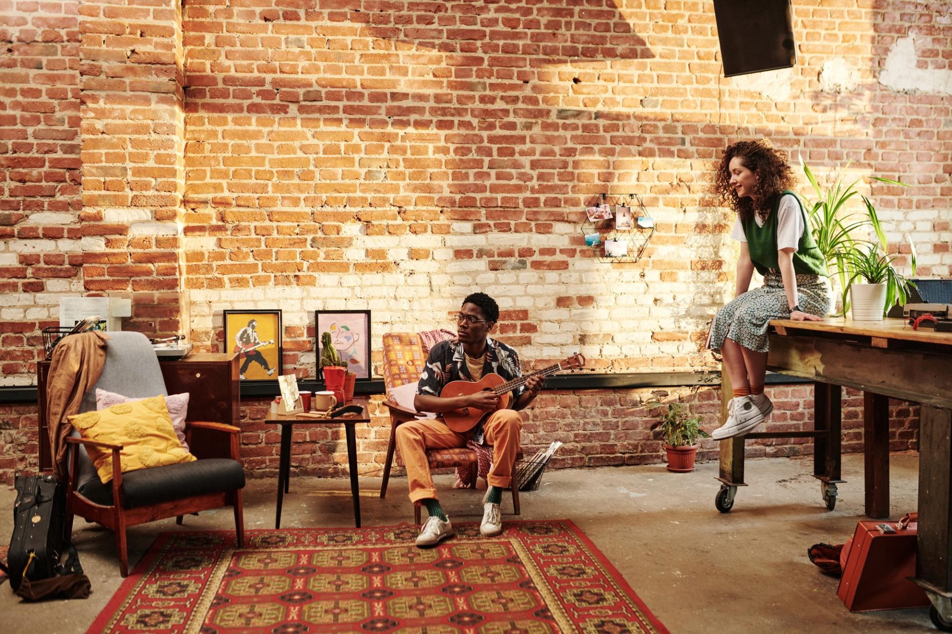 A man plays guitar on a chair in a cozy loft with exposed brick walls, while a woman sits on a wooden table nearby. Art, plants, and rugs decorate the warm, stylish space.
